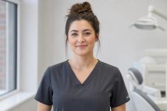 Friendly dental professional in scrubs standing in a bright dental clinic, patient chair visible.