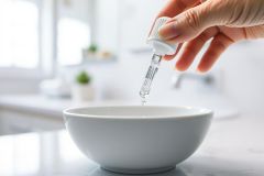 Close-up of a hand dispensing a clear liquid drop into a white bowl, likely skincare or essential oil. Clean, bright bathroom background with marble countertop and window. Focus on purity and wellness.