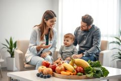 Doctor consults with a young family about healthy eating. A vibrant display of fruits and vegetables sits on a table, showcasing a focus on nutrition and wellness in a bright, modern home setting. Family health, doctor visit, healthy food.