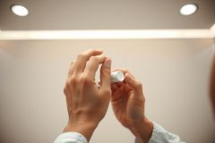Close-up of hands holding a small white container, possibly skincare or cosmetic product. Clean, bright environment with soft lighting. Focus on product and hands.