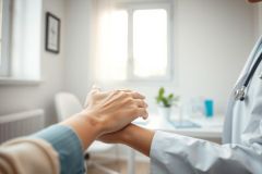 Doctor comforting patient, holding hands in a bright, modern medical office. Image conveys healthcare, support, empathy, and trust. White coat, stethoscope, and window provide context. Soft lighting and neutral colors create a calming atmosphere.