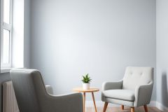 Minimalist interior design featuring two gray armchairs and a small wooden table with a potted plant. The scene is set against a light gray wall with a window, creating a calm and modern living room or reading nook aesthetic.