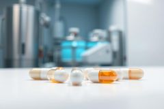 Pharmaceutical capsules in a modern laboratory setting. White and orange pills are scattered on a white surface with blurred blue and silver equipment in the background. Represents medicine, healthcare, and drug manufacturing.