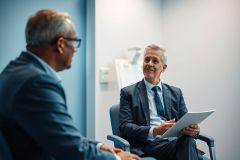 Business meeting. Two mature men in suits converse in a modern office setting. One holds a tablet. Blue and gray tones dominate the image, suggesting a professional and corporate environment. Focus on collaboration and discussion.