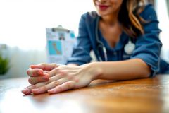 Close-up of hands gently resting on a wooden table. A woman in a blue shirt is visible in the background, with a blurred view of a building and sky. Warm lighting and a serene atmosphere.