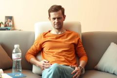 The image shows a man sitting on a couch, possibly in a living room. He is wearing an orange long-sleeved shirt and appears to be looking down with a slight frown on his face.