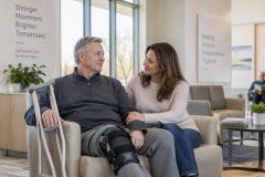Older man with knee brace and crutches seated in orthopedic clinic, companion offering support.