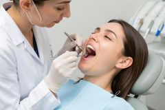 Female dentist wearing gloves examines a young woman's open mouth with mirror and probe.