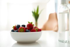 Fresh strawberries and blueberries in a white bowl on a bright surface. A glass of water and greenery are visible in the background. Healthy food, summer fruit, close-up.