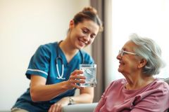 A caregiver offers water to an elderly woman in a bright, comfortable setting. The scene depicts caregiving and senior support, showcasing compassion and wellbeing.