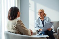 Doctor consults with a patient in a bright, modern medical office. A smiling female doctor, wearing a stethoscope, reviews paperwork while speaking with a patient. Focus on healthcare, consultation, and medical examination in a comfortable setting.