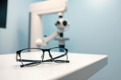 Eyeglasses resting on a white table in an optometrist's office. A blurry vision testing machine is visible in the background. Blue and white color scheme, representing eye care and vision correction services. Focus on glasses, eye exam, and optical equipment.