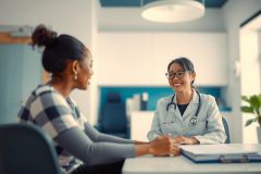 Doctor consults with a patient in a bright, modern office. The doctor wears a white coat and stethoscope, discussing health concerns with a friendly and approachable demeanor. A file folder and medical tools rest on the table.