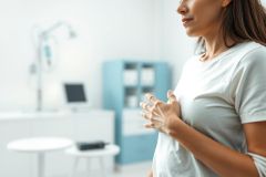 Woman experiencing chest pain or discomfort in a medical setting. Close-up shot showing her hand on her chest in a white t-shirt, with a blurred background of a doctor's office including medical equipment and a blue cabinet. Focus on health, pain, and healthcare.