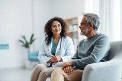 A doctor consults with a senior patient in a bright, modern medical office. The image shows a diverse healthcare setting with a female doctor in a white coat speaking with an older man. Warm tones and natural light create a comforting atmosphere, representing patient care and medical consultation.