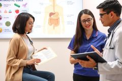 In this image, three individuals are engaged in a medical consultation. The room appears to be a modern clinic or hospital setting with educational posters and medical equipment. 