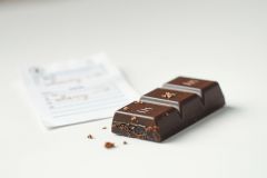 Dark chocolate bar broken into pieces on a white surface with a bakery label nearby. Rich brown tones and a blurred background create a sweet treat food photography image.
