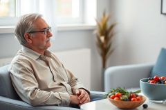 Senior man in glasses thoughtfully sitting in a modern living room. He's wearing a beige shirt and appears relaxed in a blue armchair. Bowls of fresh berries and fruit are on a nearby table, suggesting healthy living and retirement lifestyle. Bright, natural light fills the room.