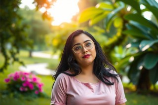 The image shows a person, likely a woman given the hairstyle and clothing. She is wearing eyeglasses and has dark hair. The individual is situated in a garden-like setting with trees and bushes in the background.