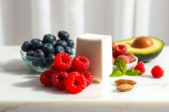 Fresh berries including raspberries, blueberries, and sliced avocado are arranged on a white surface, showcasing a vibrant and healthy food still life. The image features bright colors and natural light.