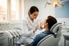 A professional female dentist in a white lab coat performs a dental examination on a male patient seated in a dental chair. Set in a bright, modern dental clinic, the scene includes specialized oral healthcare tools, an overhead examination light, and soft natural light from a window, emphasizing a clean and clinical environment.