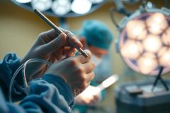 Surgical procedure in progress. Close-up view of a surgeon's hands holding surgical instruments under bright operating room lights. Medical team visible in background. Focus on precision and sterile environment.