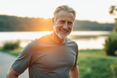 Smiling senior man with gray hair enjoys an outdoor walk near a lake at sunset. The image depicts active aging, fitness, and healthy lifestyle with warm golden light reflecting on the water and surrounding greenery. A happy, fit man enjoying nature.
