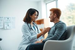 A friendly female doctor in a white coat and stethoscope examines a young man during a medical consultation in a bright, modern clinic. The scene conveys healthcare, patient care, and a positive doctor-patient relationship. Warm lighting and a clean, professional environment are prominent.