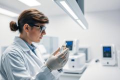 Female scientist in a lab examining a test tube. The modern laboratory setting features white walls and advanced medical equipment. Focus is on scientific research, healthcare, and laboratory analysis. Predominant colors are white and neutral, conveying a clean and professional environment.