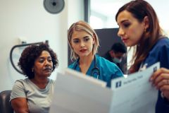A healthcare professional in scrubs reviews a patient's chart in a clinical setting. The image depicts a collaborative medical discussion, featuring a diverse group of individuals in a modern office environment.