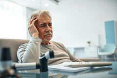Concerned senior man sitting in a modern office or meeting room, hand on head, looking thoughtful. Papers and microphone visible on the table. Neutral color palette with a focus on white and gray tones. Represents stress, worry, or decision-making.