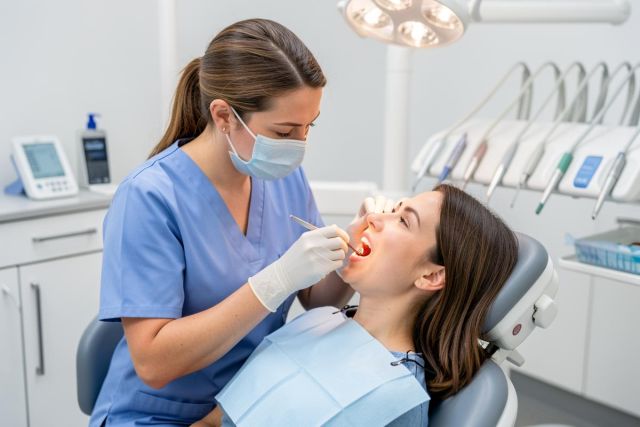 Dental hygienist wearing a mask examines a patient's open mouth during a routine dental checkup.