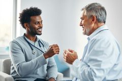 Doctor and patient consultation. A young African American doctor wearing a stethoscope discusses health concerns with an older man in a bright, modern office setting. Focus on healthcare, medical advice, and professional patient care. Blue and gray tones dominate the scene.