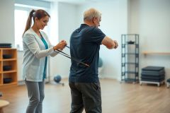 Physical therapy session: An older man exercises with the assistance of a female therapist in a bright, modern gym. The scene depicts rehabilitation and senior fitness, utilizing resistance bands for strength training. Focus on recovery and healthcare.