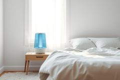 Modern bedroom scene featuring a blue humidifier on a wooden nightstand next to a neatly made white bed. Soft, natural light streams through a window, creating a calming and minimalist atmosphere. Ideal for home decor, bedroom design, or humidifier product imagery.