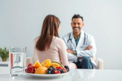 Doctor consult with patient about healthy eating. Image shows a doctor in a white coat discussing nutrition with a woman, with a plate of fresh fruits like oranges, berries, and a glass of water on a table.