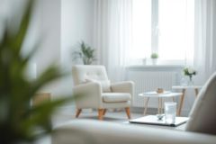 Bright, airy living room interior featuring a cream armchair, small wooden side tables, and natural light streaming through a window. The space showcases a minimalist, Scandinavian design with soft white walls and touches of greenery. A cozy and inviting home decor scene.