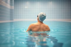 Rear view of a swimmer wearing a swim cap in a blue indoor pool. The scene features cool blue tones and a tranquil aquatic environment, suggesting fitness, training, or recreational swimming. Focus is on the water and the swimmer's back.