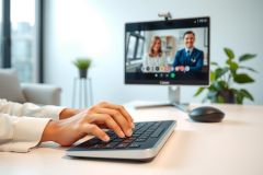 Close-up of hands typing on a keyboard in a modern office setting. A video call is visible on the monitor, featuring two people. Bright and airy workspace with a clean aesthetic. Focus on technology and remote communication.