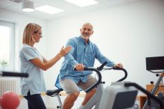 Senior man exercises on a stationary bike with a physical therapist assisting. Healthcare, rehabilitation, and fitness theme in a bright, modern clinic setting.