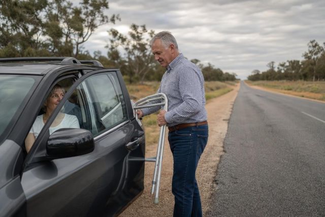 Older man helping woman with a walker at car on rural roadside, travel context.