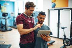 Fitness trainer with tablet reviewing workout plan with client in gym. Red shirt, blue gym background, focus on health and exercise.