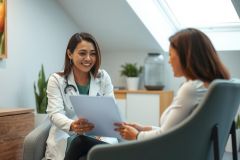Doctor in a white coat consults with a patient in a bright, modern office. The scene conveys healthcare, consultation, and professional medical care. Green and white tones dominate the environment.