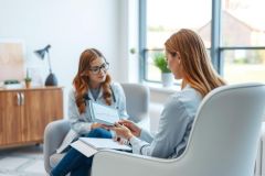 A therapist or counselor sits with a client in a modern office setting. The scene features a woman holding a chart, suggesting a consultation or discussion. The warm lighting and neutral colors create a calming atmosphere.