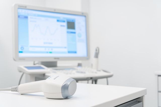 Professional medical ultrasound equipment in a bright, sterile clinic setting. A white ultrasound transducer sits in the foreground on a white table, with a blurred medical monitor displaying diagnostic data in the background. The clean, high-key lighting and blue-toned display highlight modern healthcare technology and diagnostic imaging.