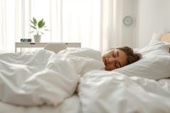 Young woman sleeping peacefully in a cozy, bright bedroom. White bedding, soft light, and a calming atmosphere create a restful scene. Focus on sleep, relaxation, and a comfortable bedroom interior. Minimalist decor with a plant adds to the serene setting.