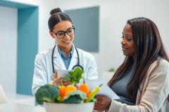 Doctor consults with a patient about nutrition in a bright, modern clinic. The image features a female doctor in a white coat reviewing documents with a patient, with a bowl of fresh vegetables (broccoli, peppers) on the table. Focuses on healthcare, wellness, and healthy eating.