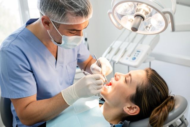 Dentist in mask and scrubs examining a reclining patient’s open mouth under an overhead dental light.