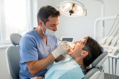 Dentist in mask and gloves examining a patient's open mouth in a dental chair under an exam light.