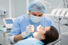 Dentist in surgical cap and mask examining a patient's open mouth with dental instruments, conveying tooth-preserving care.